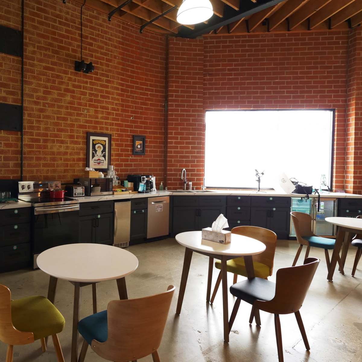 A modern kitchen area featuring stainless steel appliances, black cabinets, a large window, and several tables with colorful chairs.