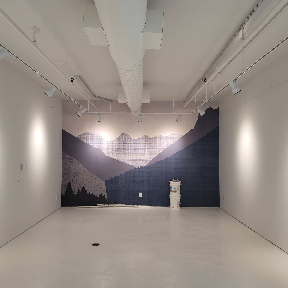 An empty gallery space features a large mural of mountains on one wall, with bright white walls and a concrete floor.