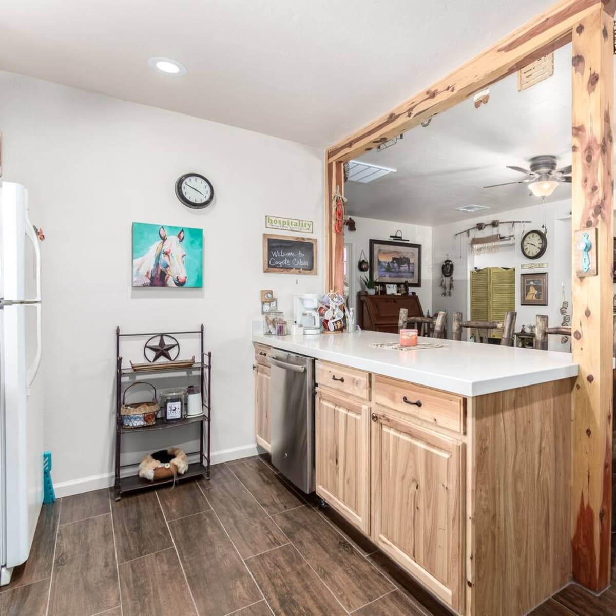 A modern kitchen with wooden cabinetry and a countertop dividing it from a dining area featuring rustic furniture and decorative artwork.