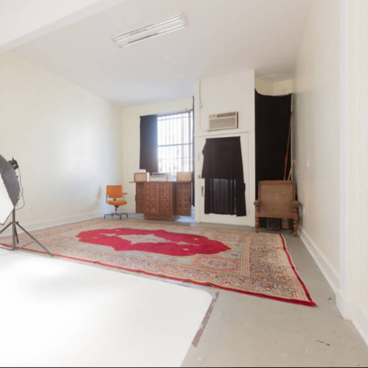 A sparse studio space with a red patterned rug, a wooden desk, an orange chair, and a large softbox light set up on the floor.