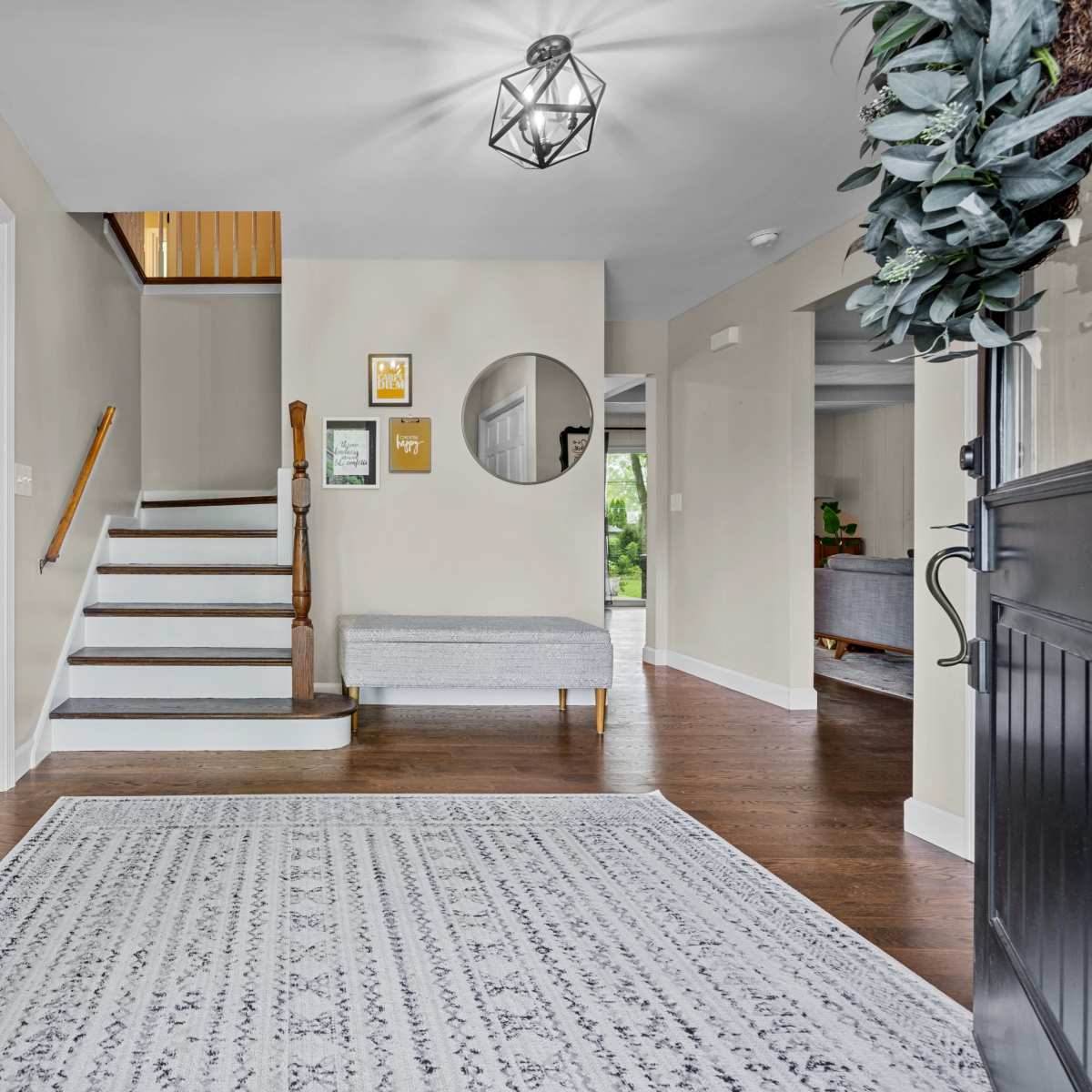 A welcoming entryway with two black doors open to reveal a staircase leading to an upper floor, framed by greenery and a decorative round mirror on the wall.