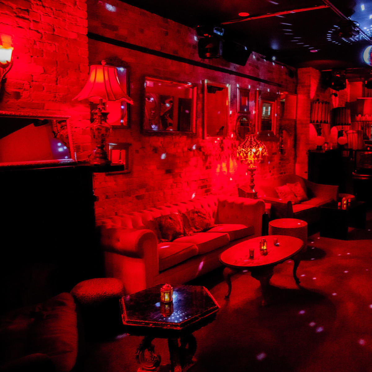 An illuminated lounge filled with red lighting, featuring a vintage sofa, tables, and decor, along with a disco ball reflecting light across the room.