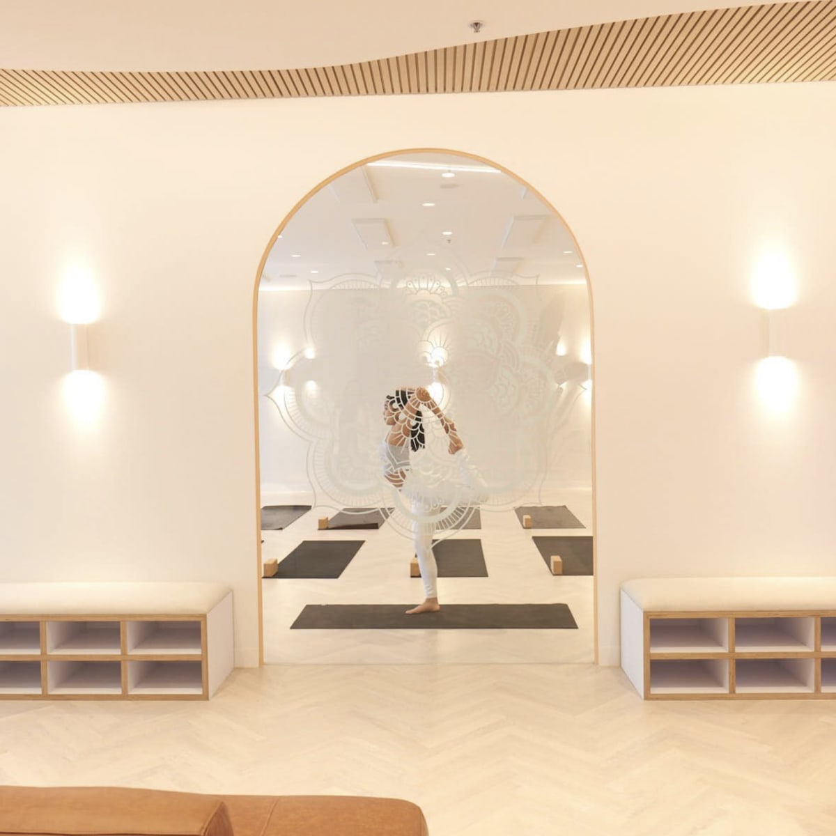 A person is practicing yoga in a minimalist studio with wooden benches and mirrored walls.