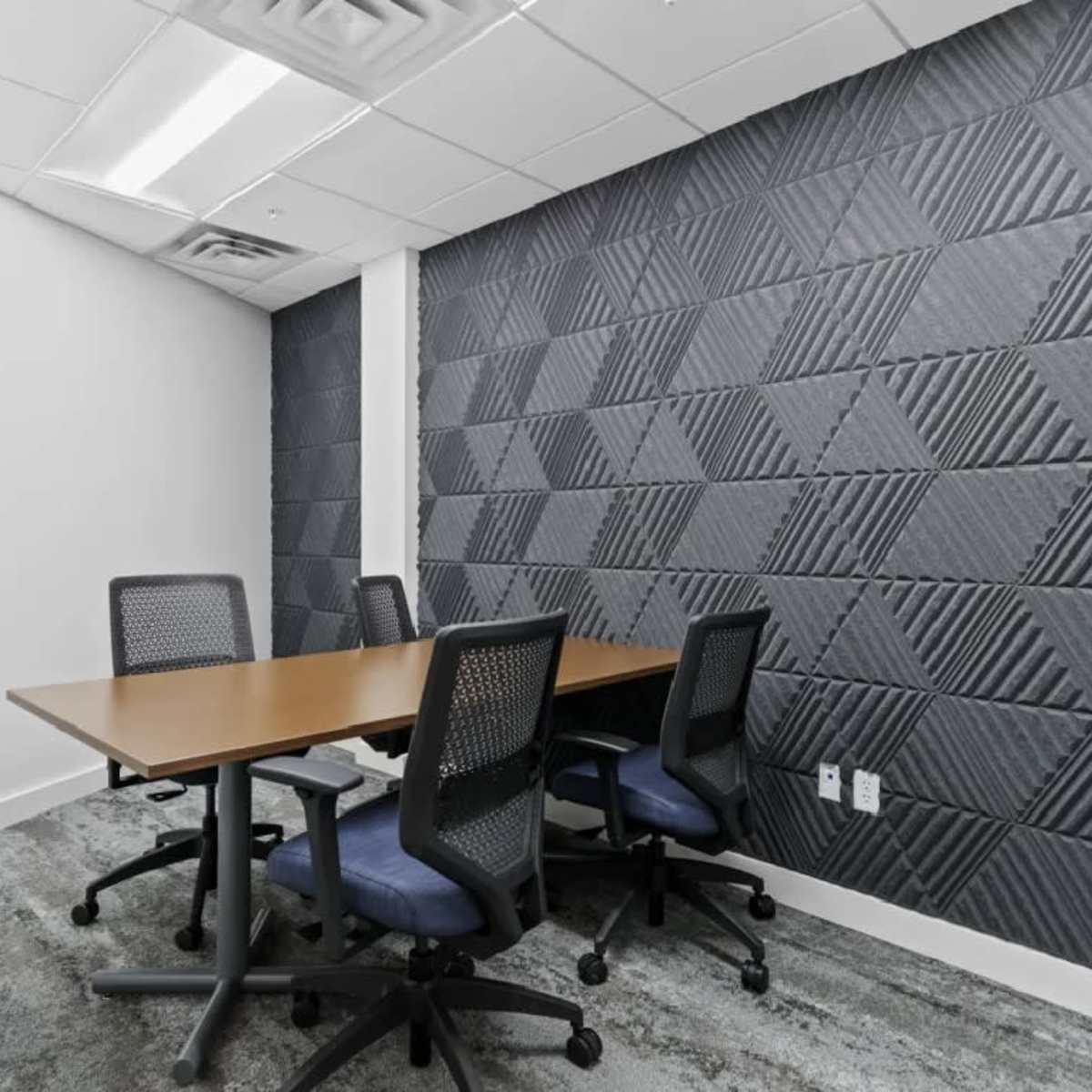 A small conference room features a wooden table surrounded by four office chairs, with textured gray walls and carpeted flooring.