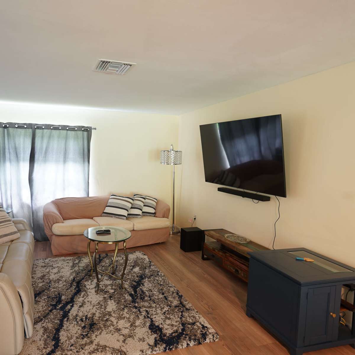 A living room with a beige couch, a light-colored recliner, a glass coffee table, and a mounted television on the wall, accompanied by muted curtains and a wooden side table.