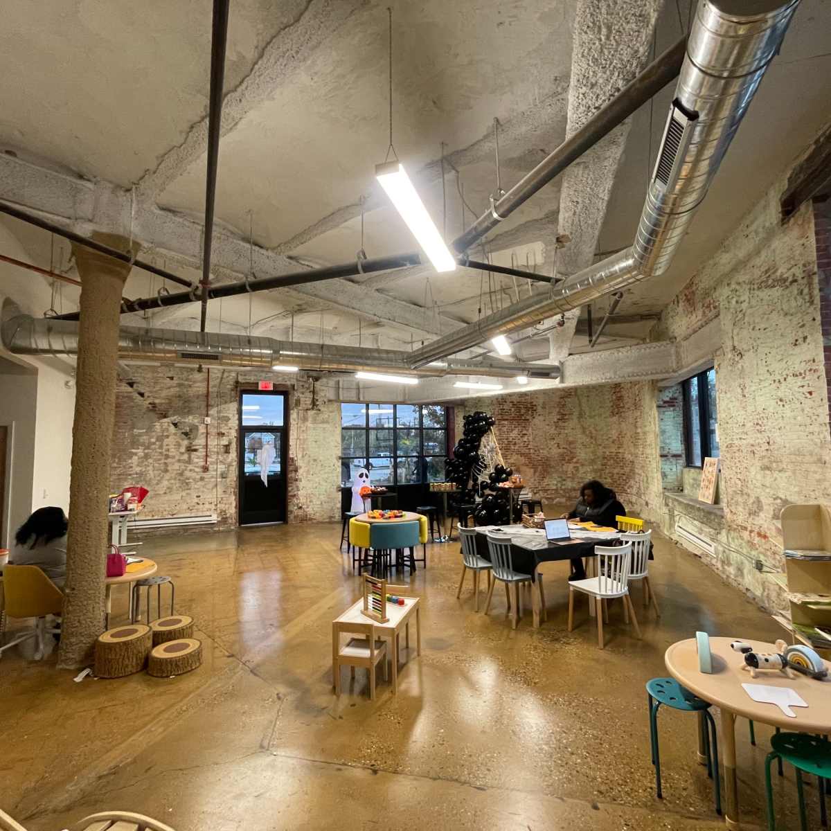 An open play area with various child-sized tables and chairs, exposed brick walls, and large windows providing natural light.
