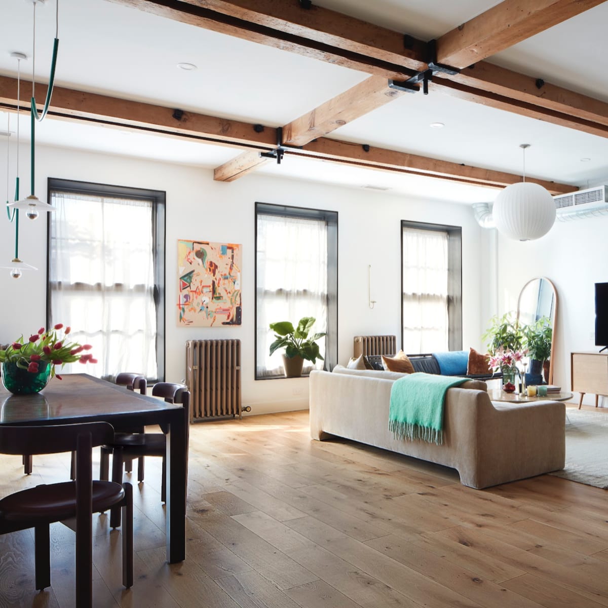 A spacious living room with a light-colored sofa, a TV on a wooden stand, large windows, and plants, all featuring wooden beams on the ceiling.