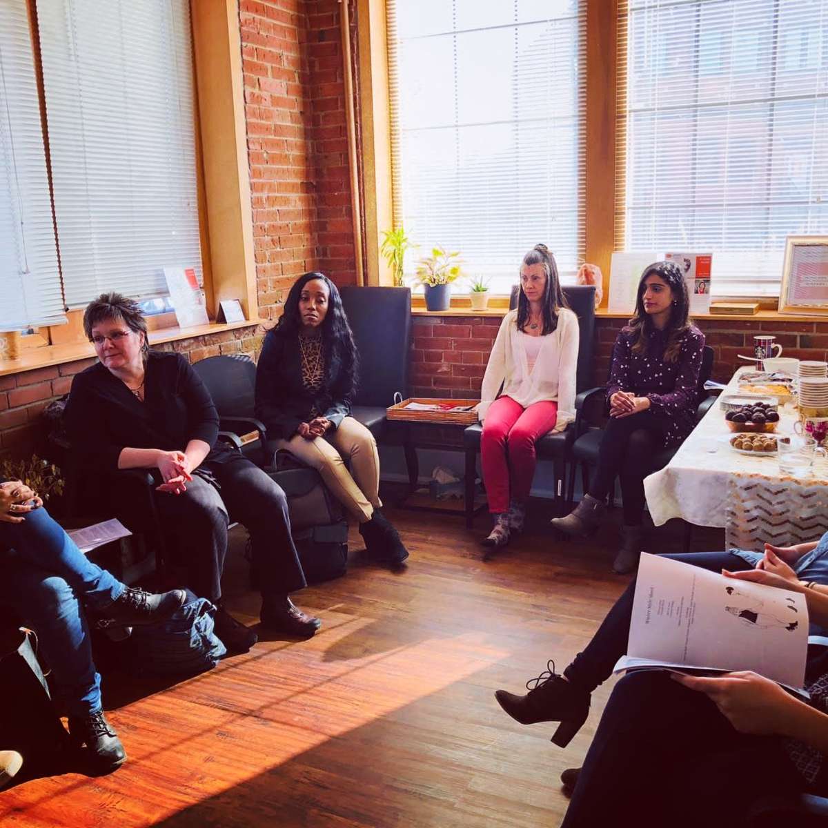 A group of five women is seated in a sunlit room with brick walls, engaging in a discussion while a table with snacks is in the background.