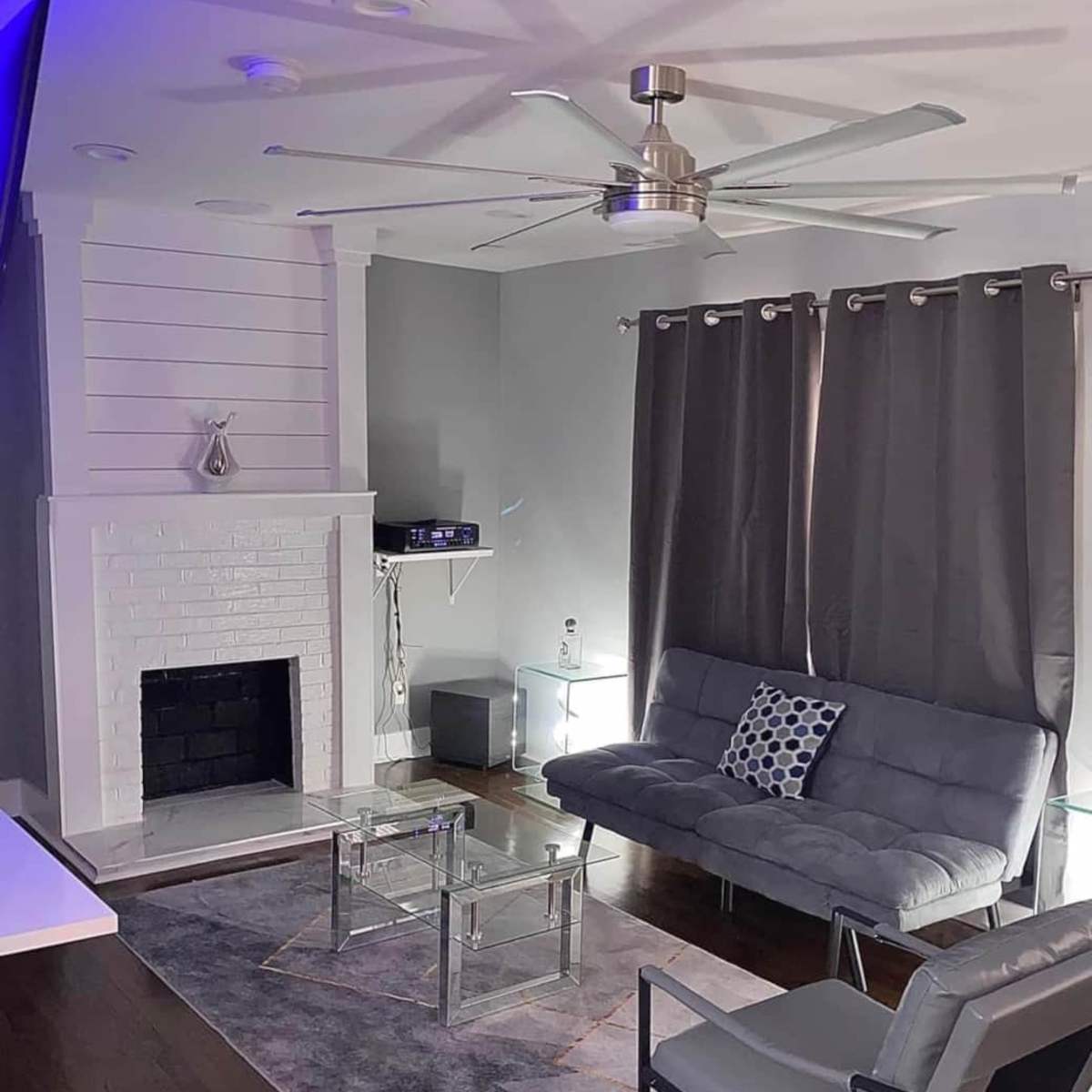 A modern living room featuring a gray sofa, a glass coffee table, a ceiling fan, and dark curtains, with a fireplace and wall-mounted shelves in the background.