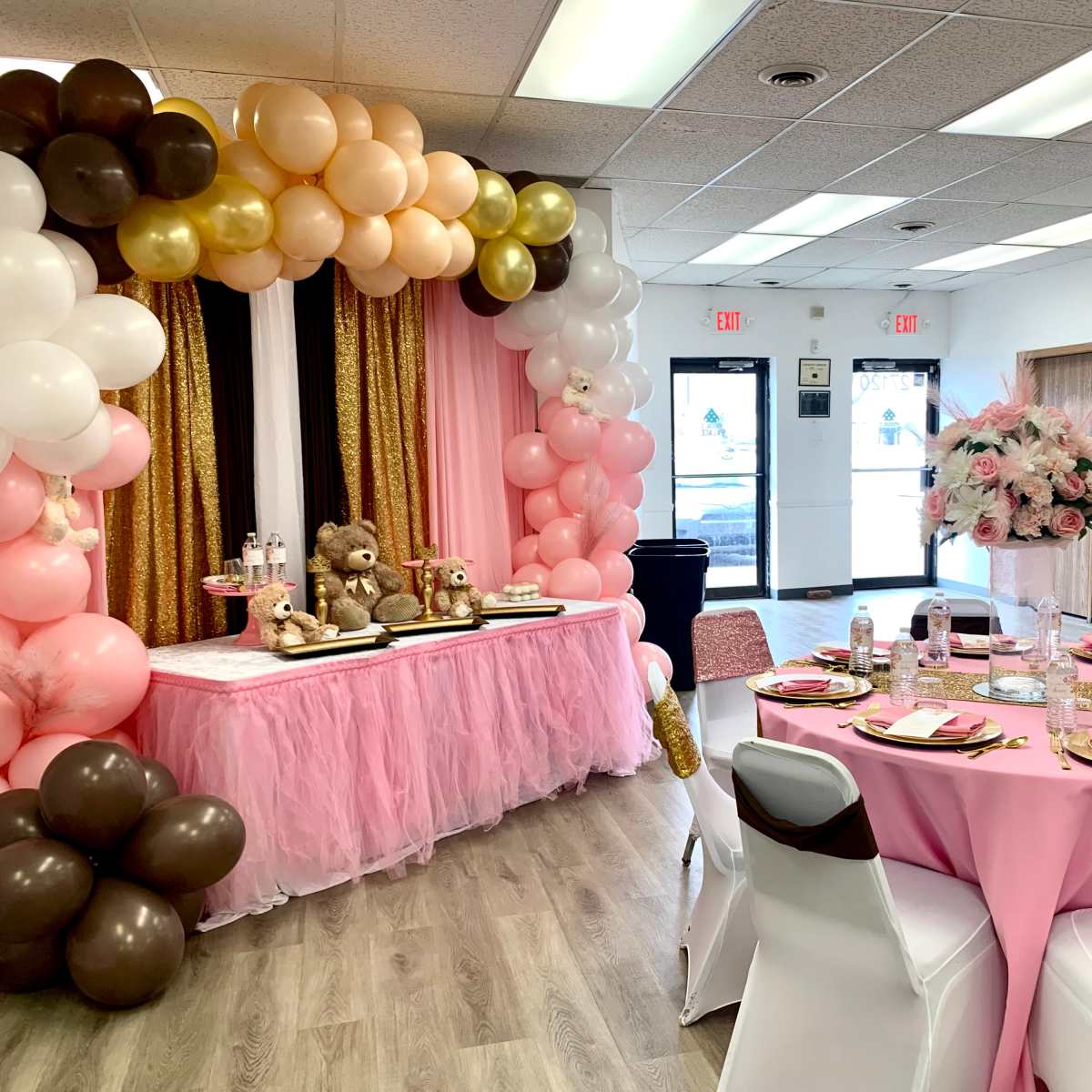 A decorated event space with pink and gold balloons, a table set for a celebration, and floral centerpieces.