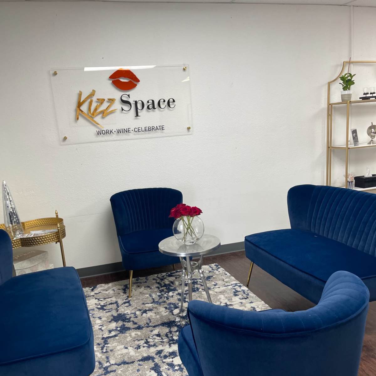 A modern lounge area with blue velvet chairs arranged around a glass table on a patterned rug, featuring a sign for "Kizz Space" on the wall.