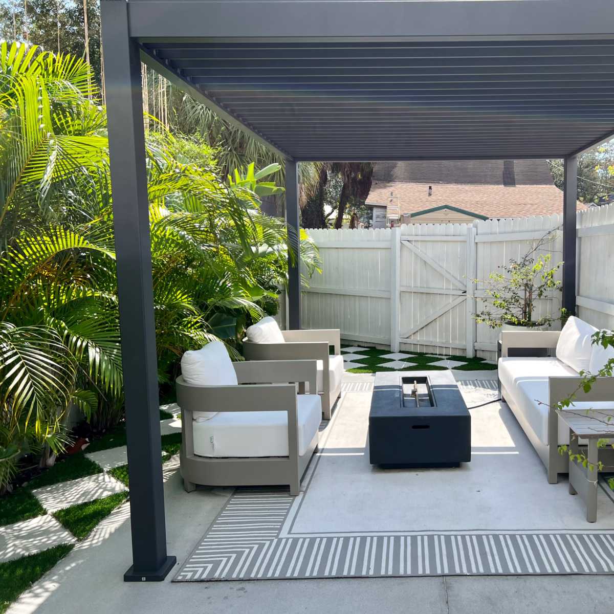 A covered outdoor seating area features a fire pit, two armchairs, and a loveseat surrounded by greenery and a patterned rug.