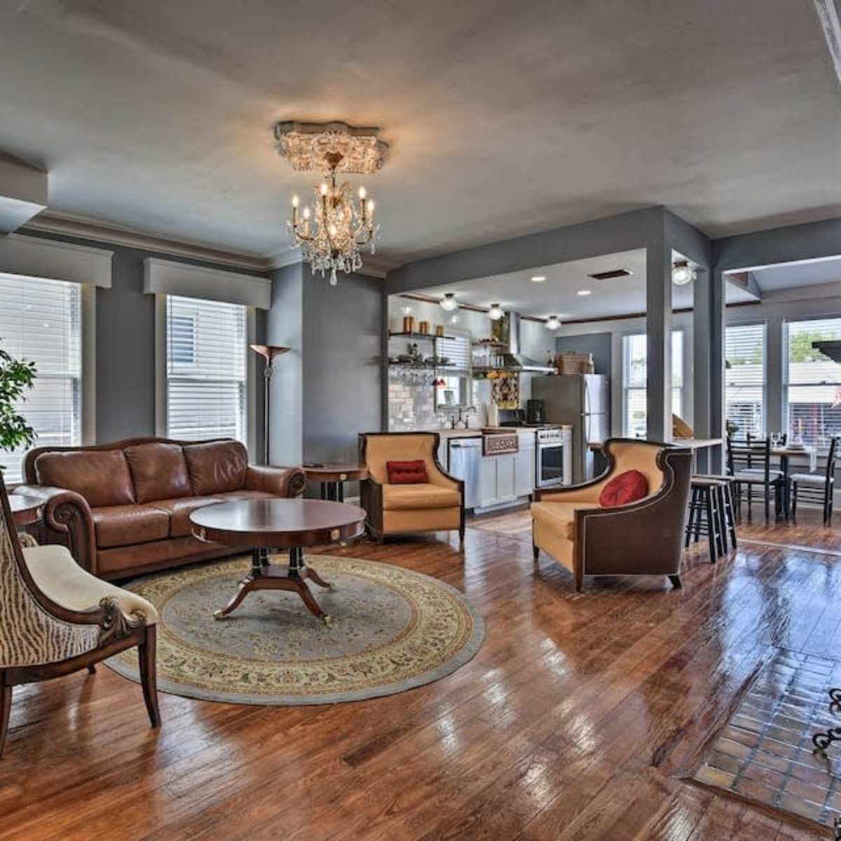 A spacious living room with hardwood floors, a brown leather sofa, and a round wooden coffee table, surrounded by windows and a kitchen area in the background.