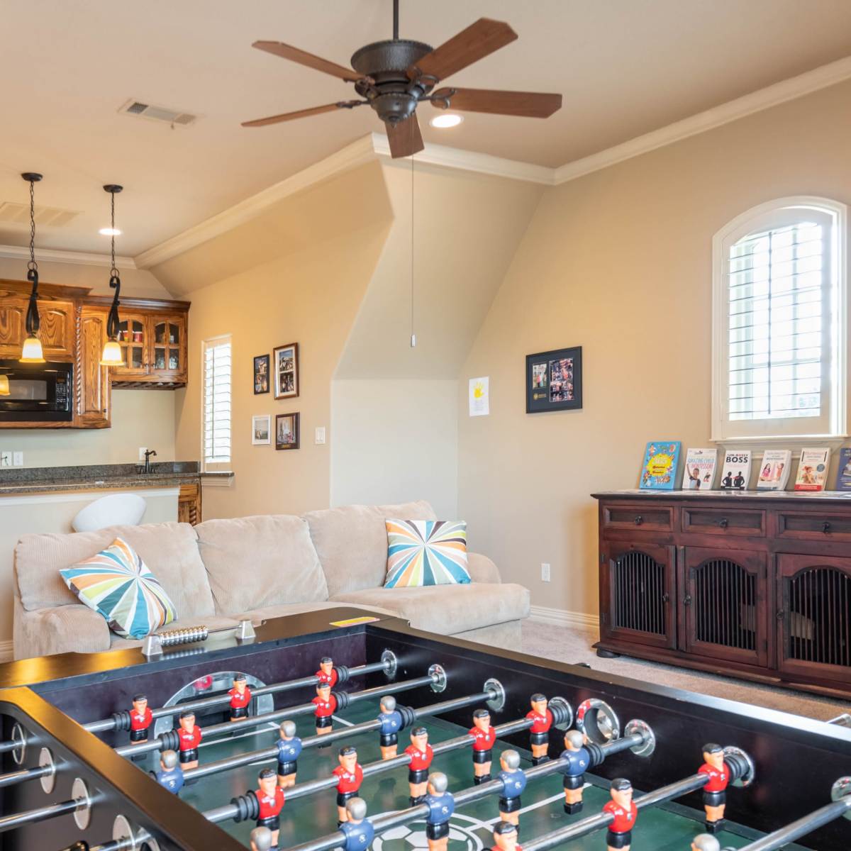 A game room featuring a foosball table, a beige couch with colorful cushions, and a kitchenette in the background.