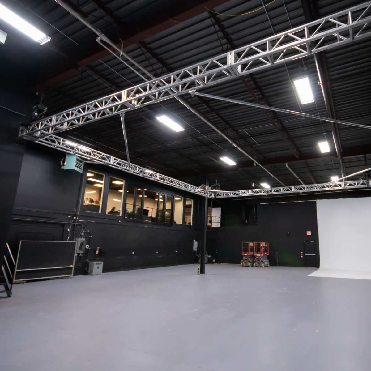 An empty photography studio with black walls, a gray floor, and a white backdrop at one end.