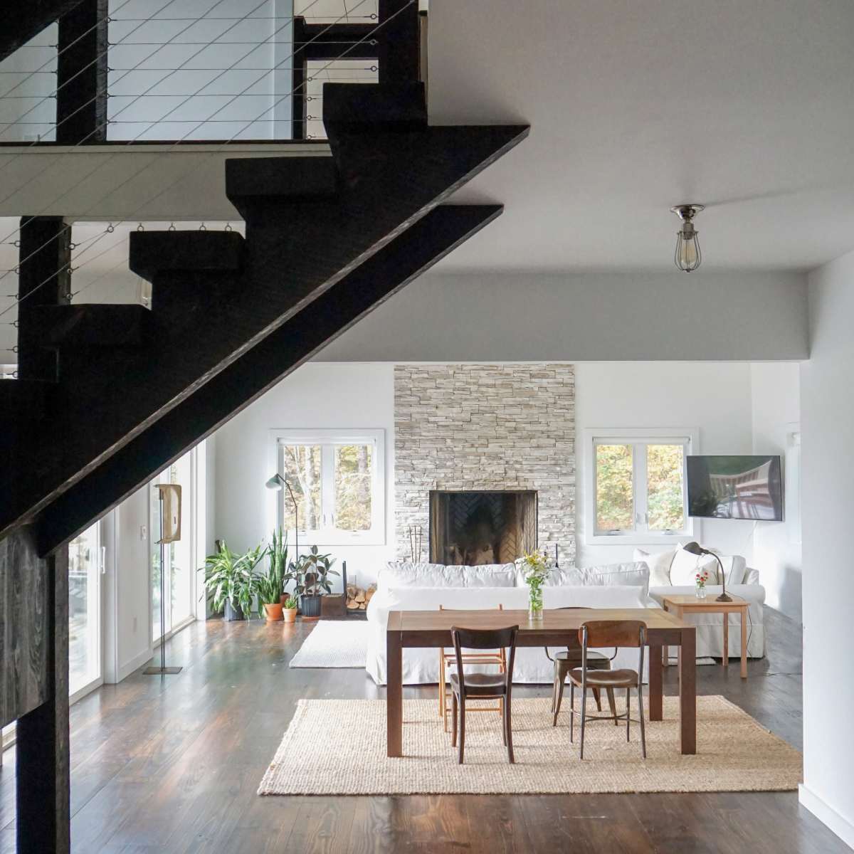A modern living room features a stone fireplace, dining area with a wooden table, and large windows allowing natural light.