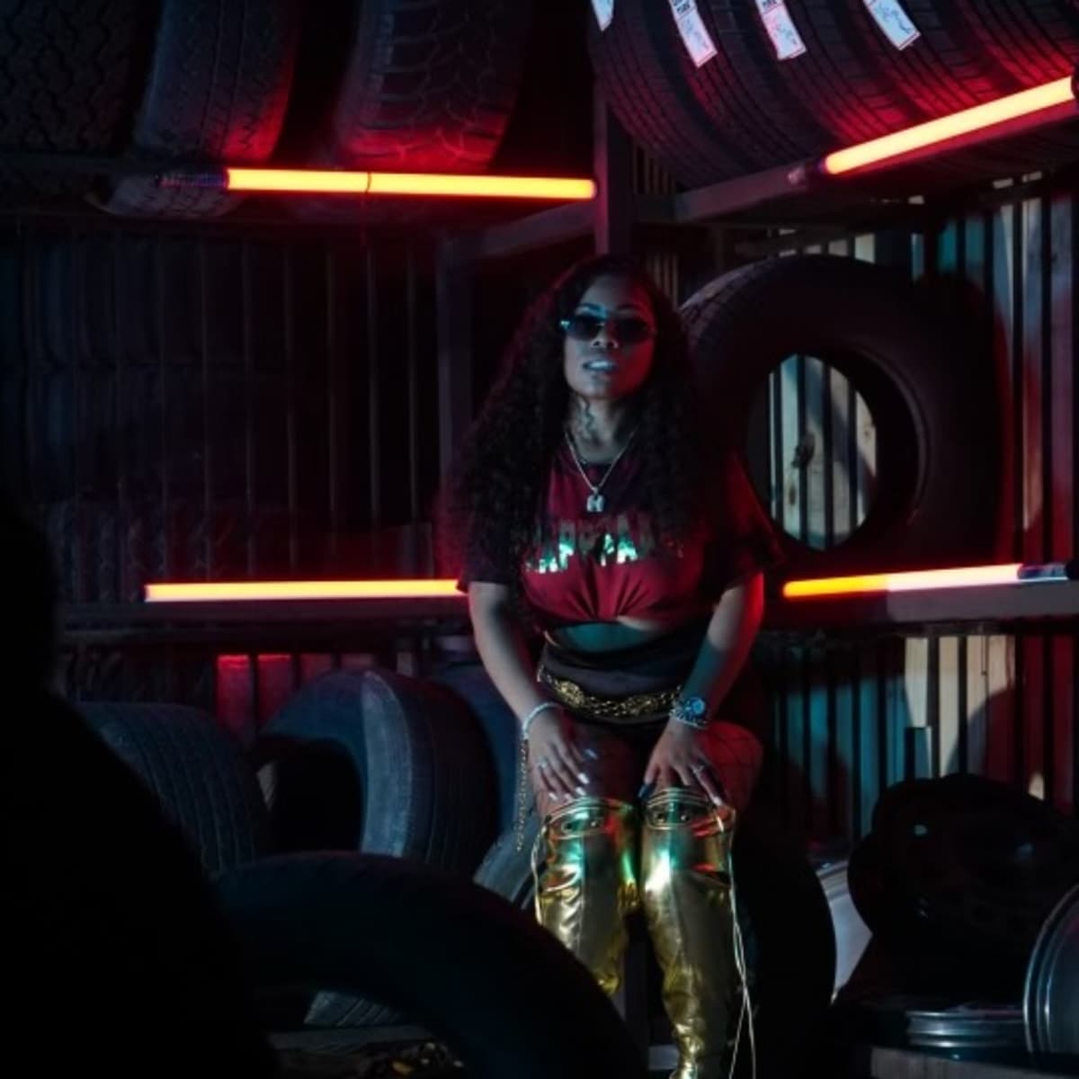A person sits on a tire in a dimly lit environment with stacked tires and neon lights in the background.
