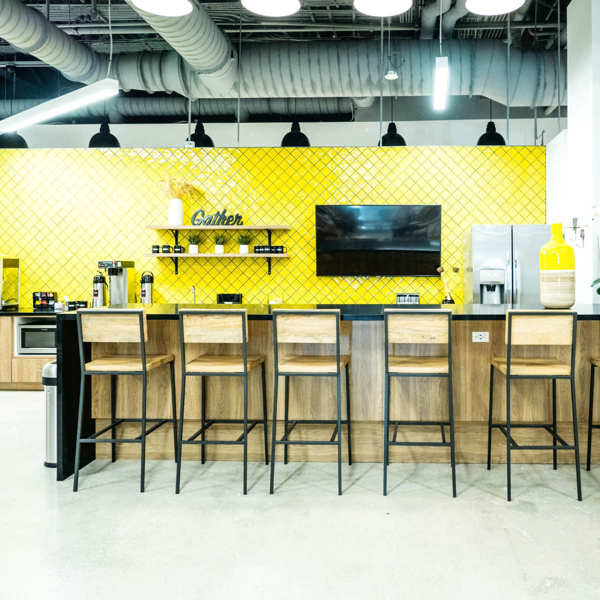 A modern kitchen with a yellow tiled wall, a large screen, and an island bar with high wooden stools arranged in front.