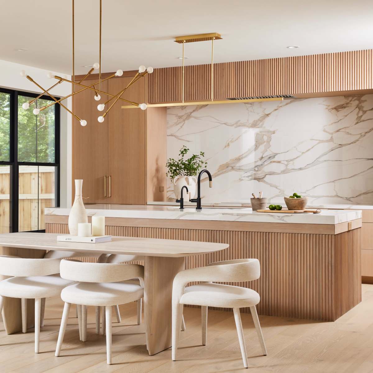 A modern kitchen features a wooden dining table surrounded by upholstered chairs, a minimalist chandelier, and a marble accent wall with a spacious island.