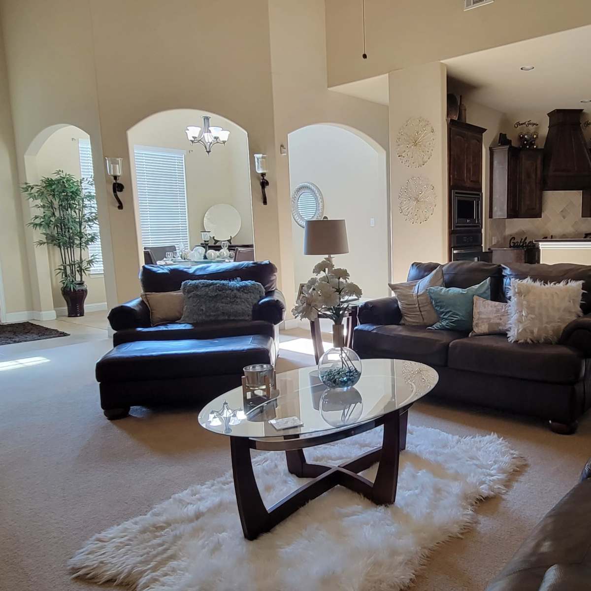 A spacious living room furnished with dark brown sofas, a glass coffee table, and decorative elements, featuring a large window and light-colored walls.