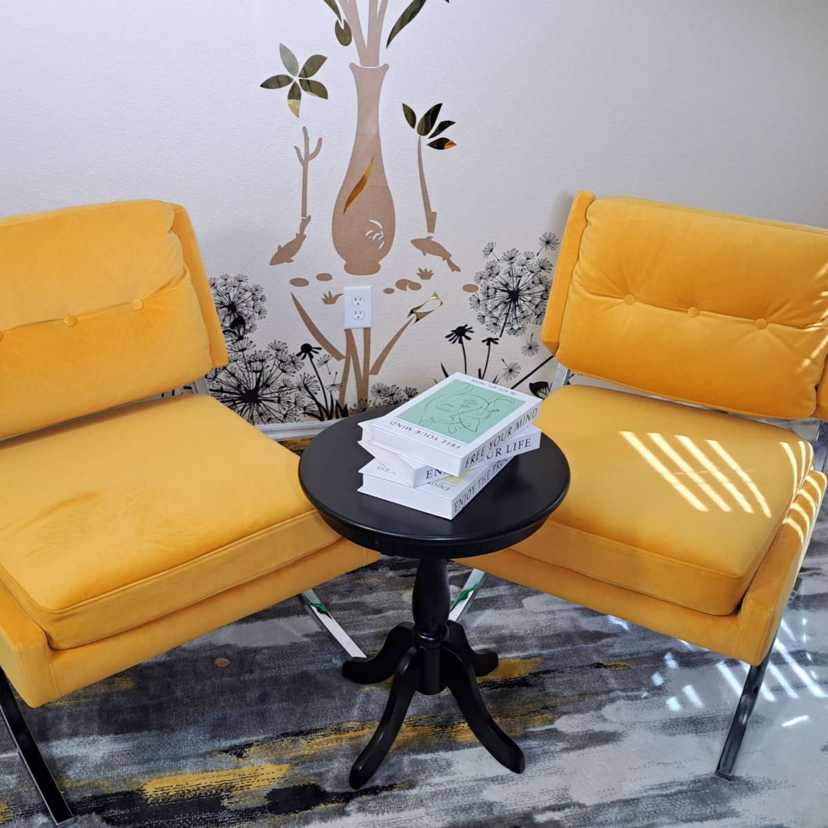 Two yellow chairs facing each other around a black circular side table, with a stack of books on top, set against a wall featuring a decorative design.