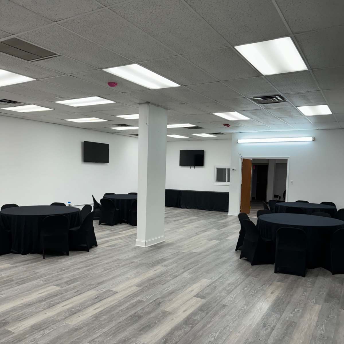 A spacious, empty room with several round tables covered in black tablecloths and TV screens mounted on the walls.