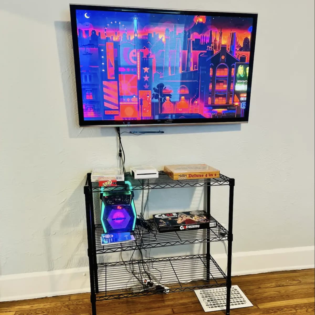 A black wire shelving unit holds a game console and magazines beneath a mounted television displaying a colorful urban skyline.