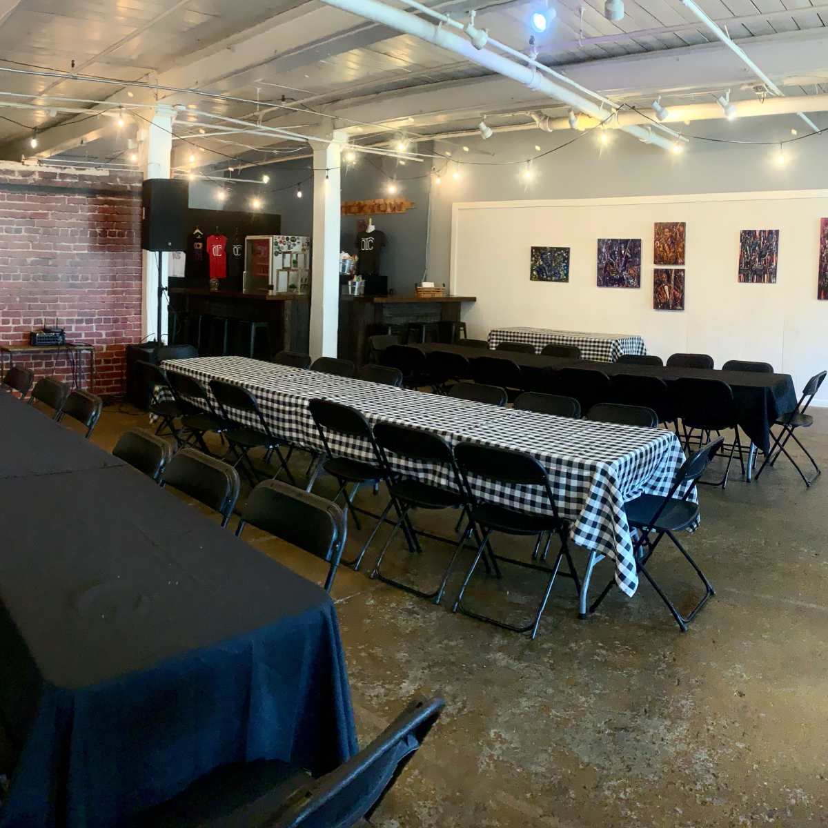 A spacious event venue with several long tables arranged in rows, a brick wall, and a bar area in the background.