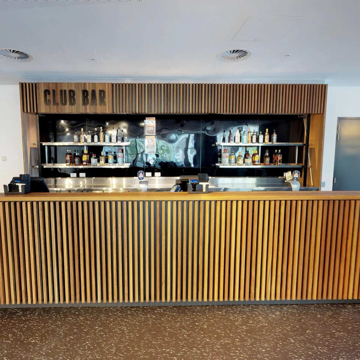 A modern bar area with a wooden slatted counter, shelves stocked with various bottles, and a minimalist design.