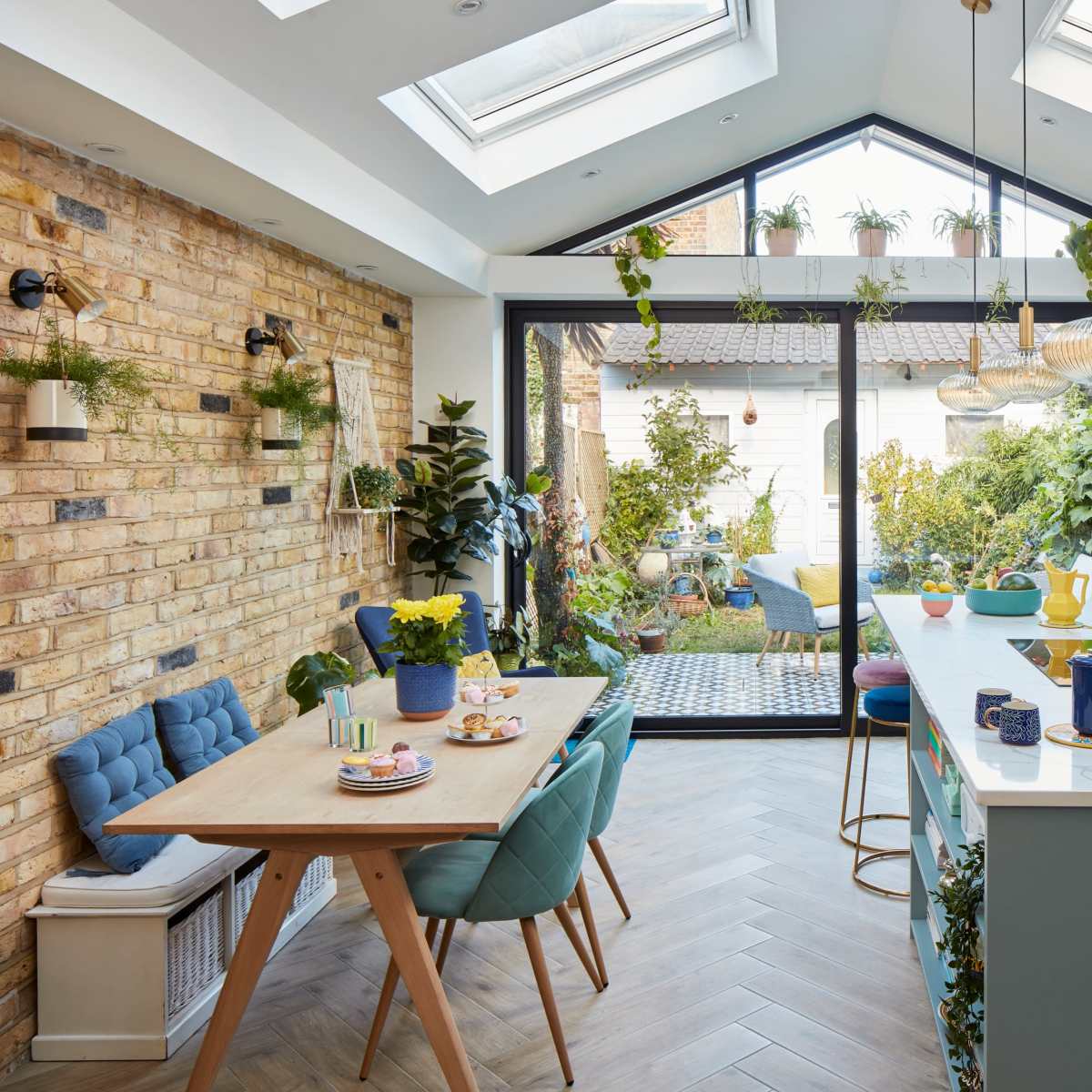 A modern kitchen with a large island, blue cabinetry, a dining table set for a meal, and abundant greenery seen through large glass doors.