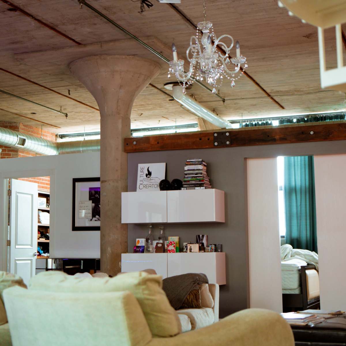 A spacious loft-style living area features a large concrete pillar and white furniture, with modern shelving units and a glimpse of a bedroom in the background.