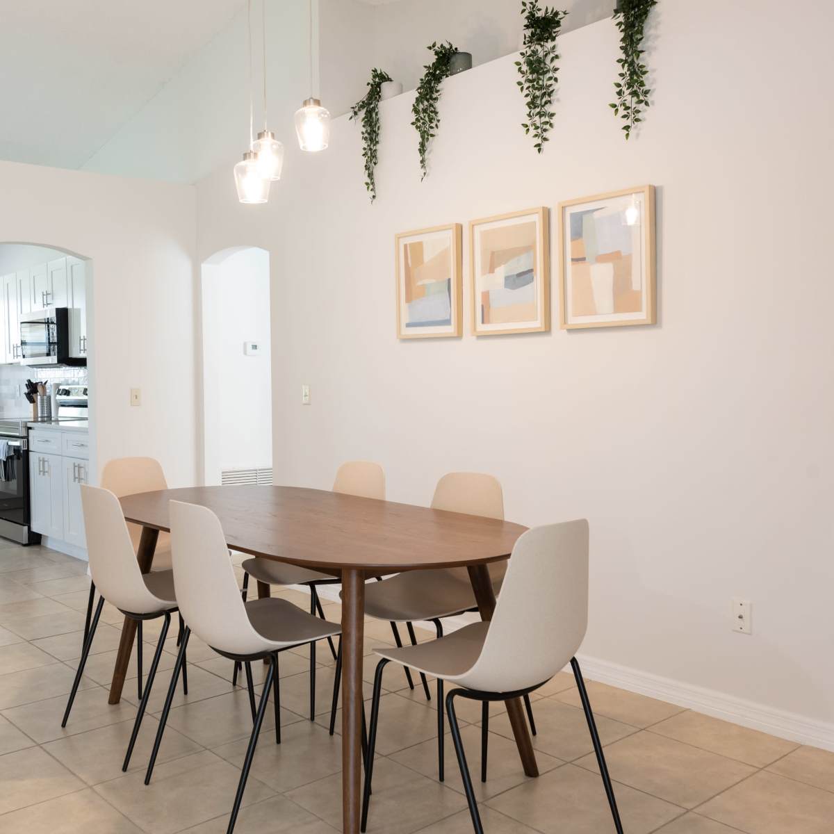 A dining area features a wooden table surrounded by six chairs, with three framed artworks on the wall and hanging plants above.