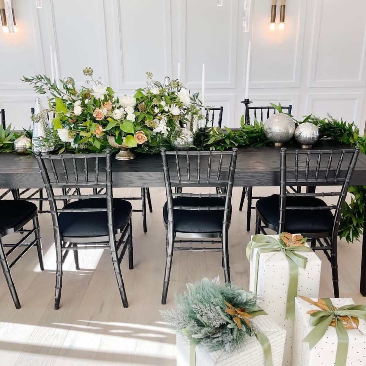 A long dining table decorated with floral arrangements, candles, and greenery, flanked by black chairs and adorned with wrapped gifts at the ends.