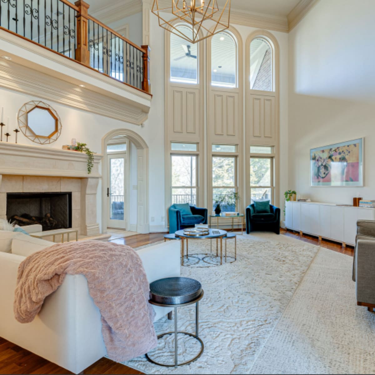 A spacious living room with high ceilings, a stone fireplace, large windows, and modern furniture arranged around a central area rug.