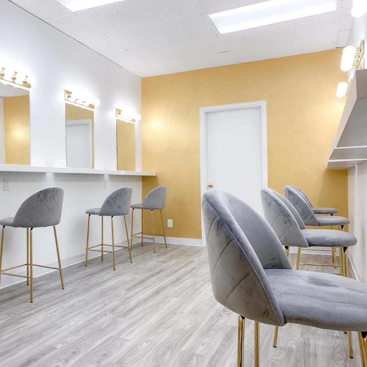 A makeup station features several gray bar stools with gold legs arranged in front of illuminated mirrors against a gold-accented wall.
