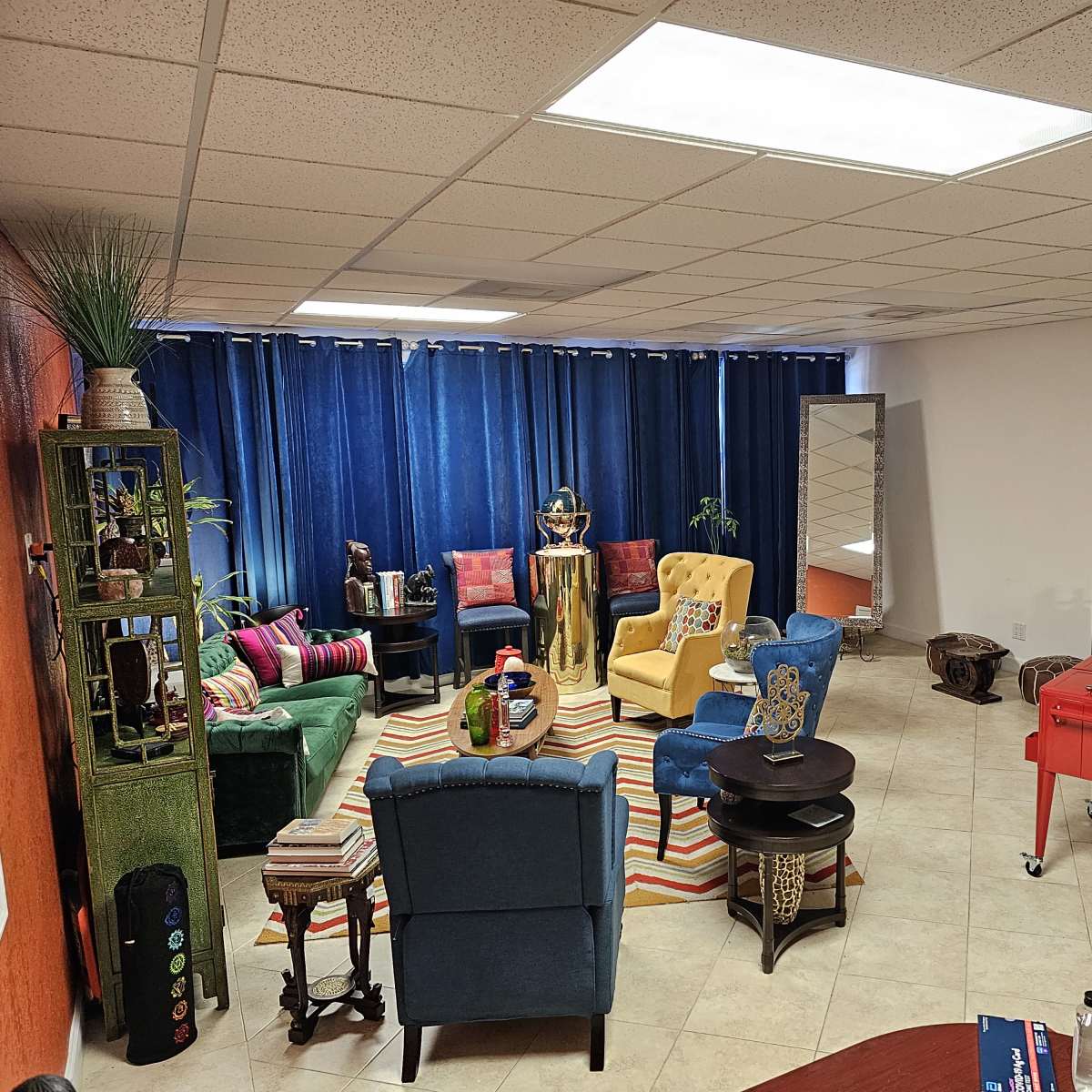 A vibrant room features colorful furniture, including a yellow and blue chair, a striped rug, and a bar cart, with large blue curtains covering a window.
