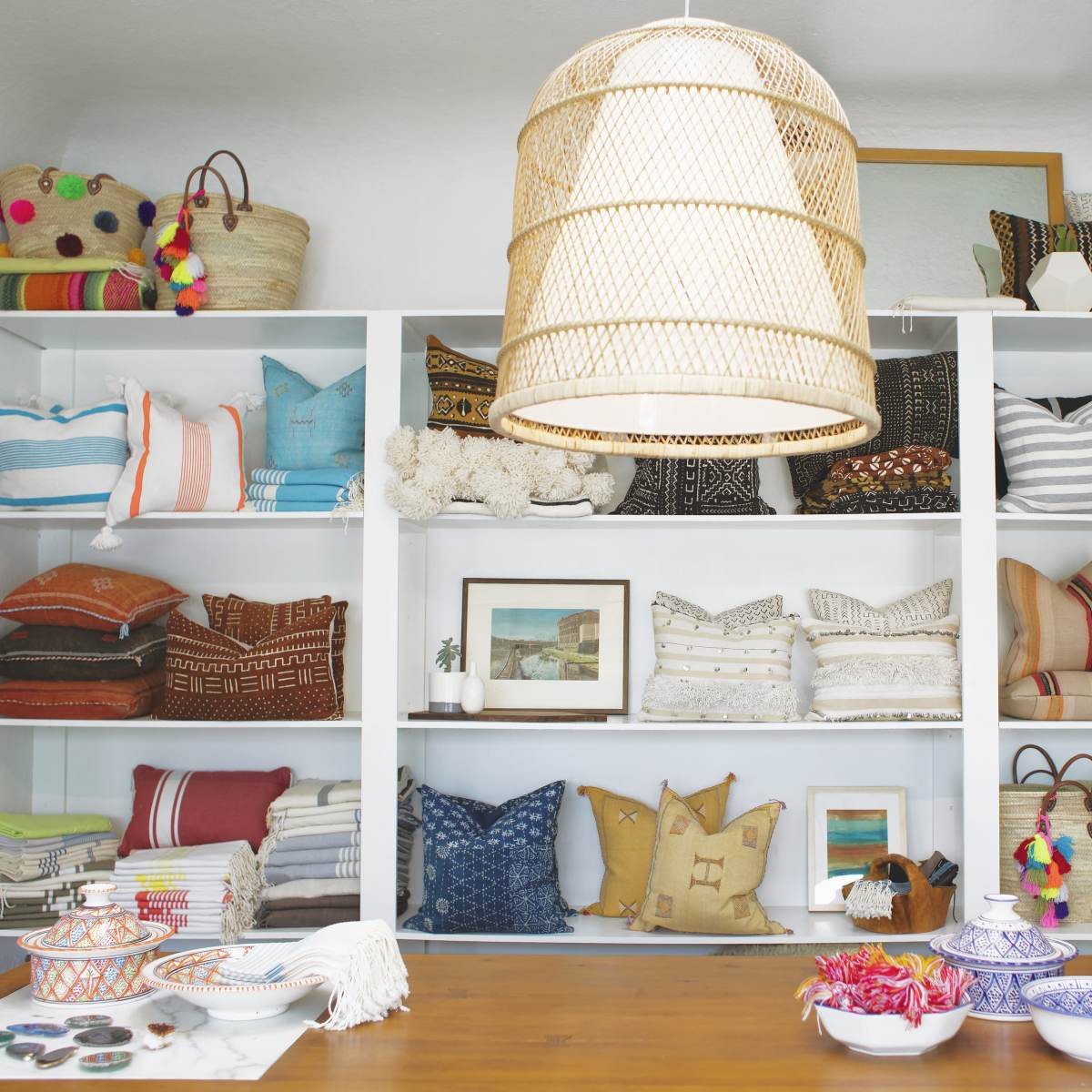 A display of various patterned pillows and woven baskets is arranged on shelves, under a large woven light fixture, in a brightly lit interior space.
