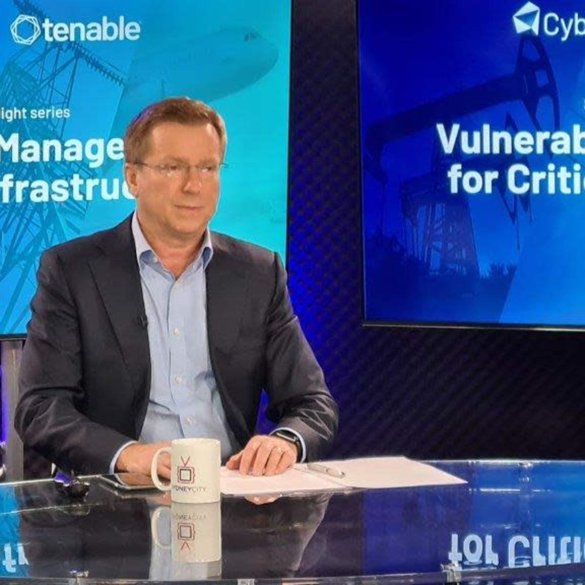 Three professionals sit at a table, discussing vulnerability management for critical infrastructure, with a large screen displaying the topic in the background.