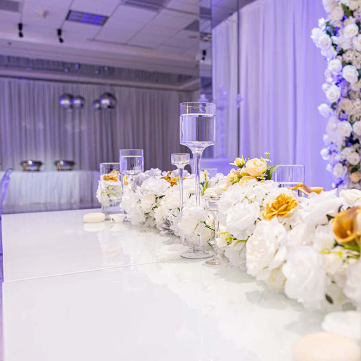 A beautifully arranged table features a floral centerpiece with white and gold flowers, surrounded by clear glasses and elegant decor in a softly lit event space.