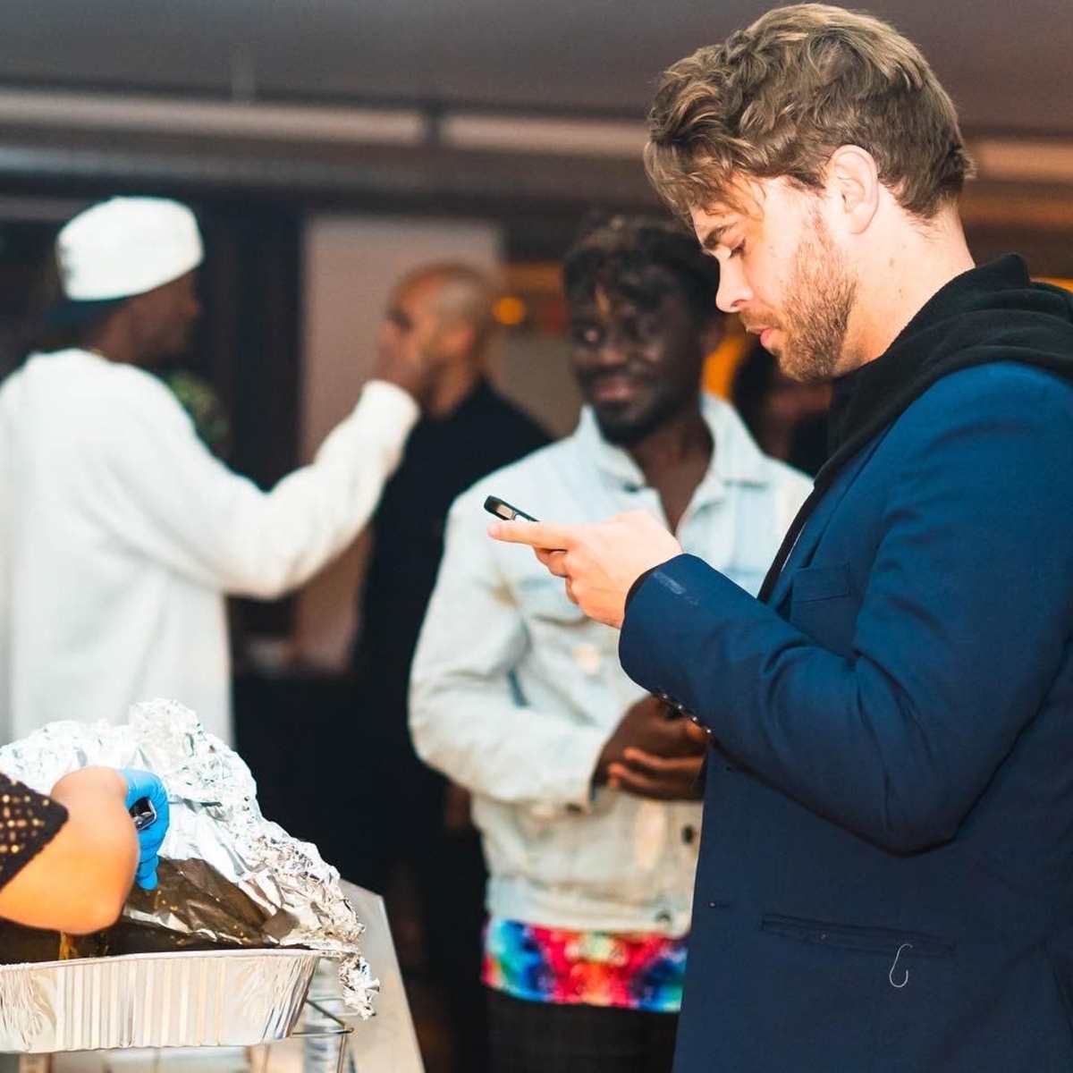 A group of people gather at an event while one person serves food from a tray and another checks their phone.