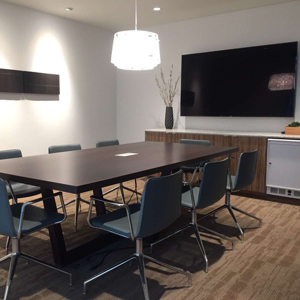 A conference room features a large rectangular table surrounded by six blue chairs, a wall-mounted television, and minimal decorative elements.