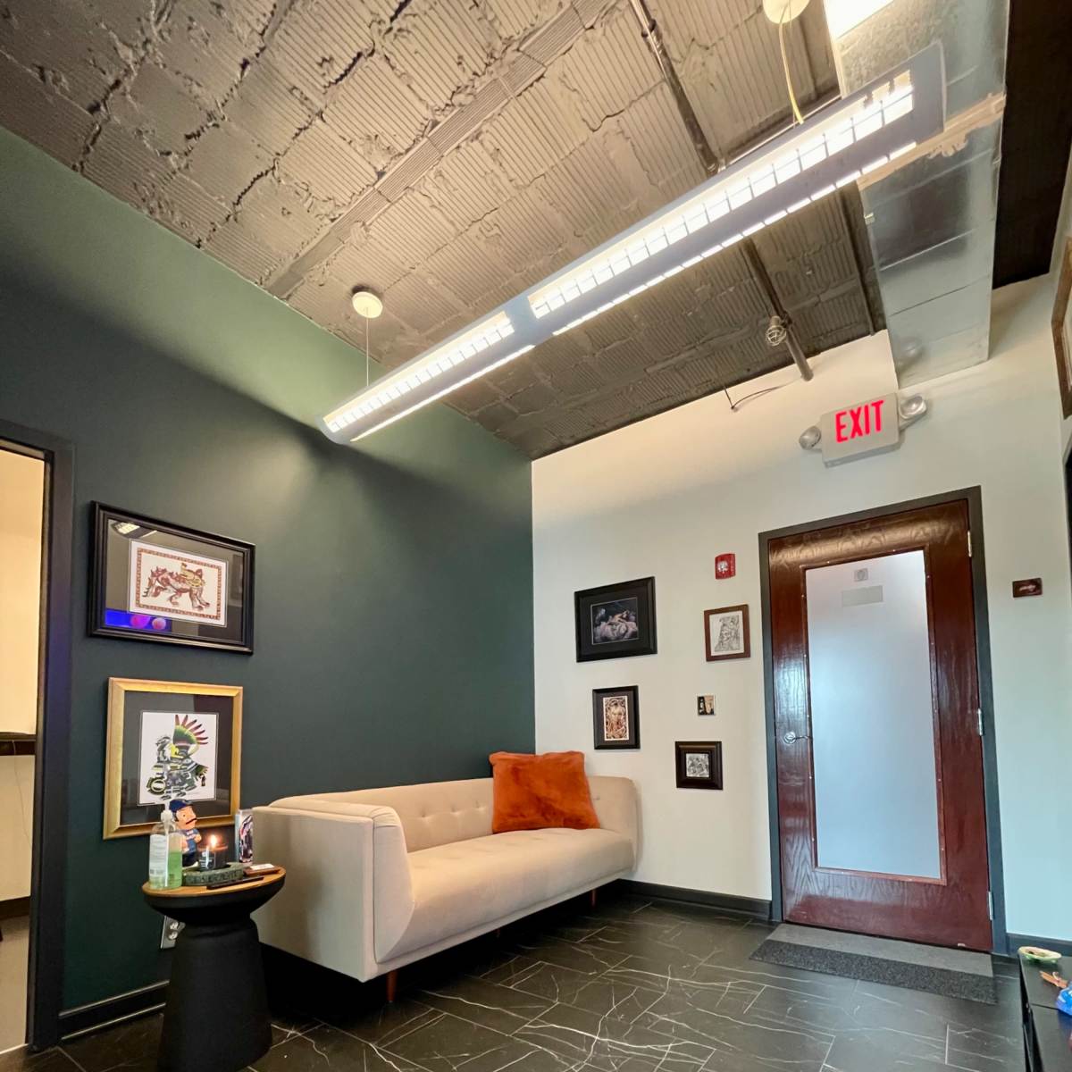 A modern waiting area with a gray sofa, dark green walls, and framed artwork, leading to a door marked with an exit sign.