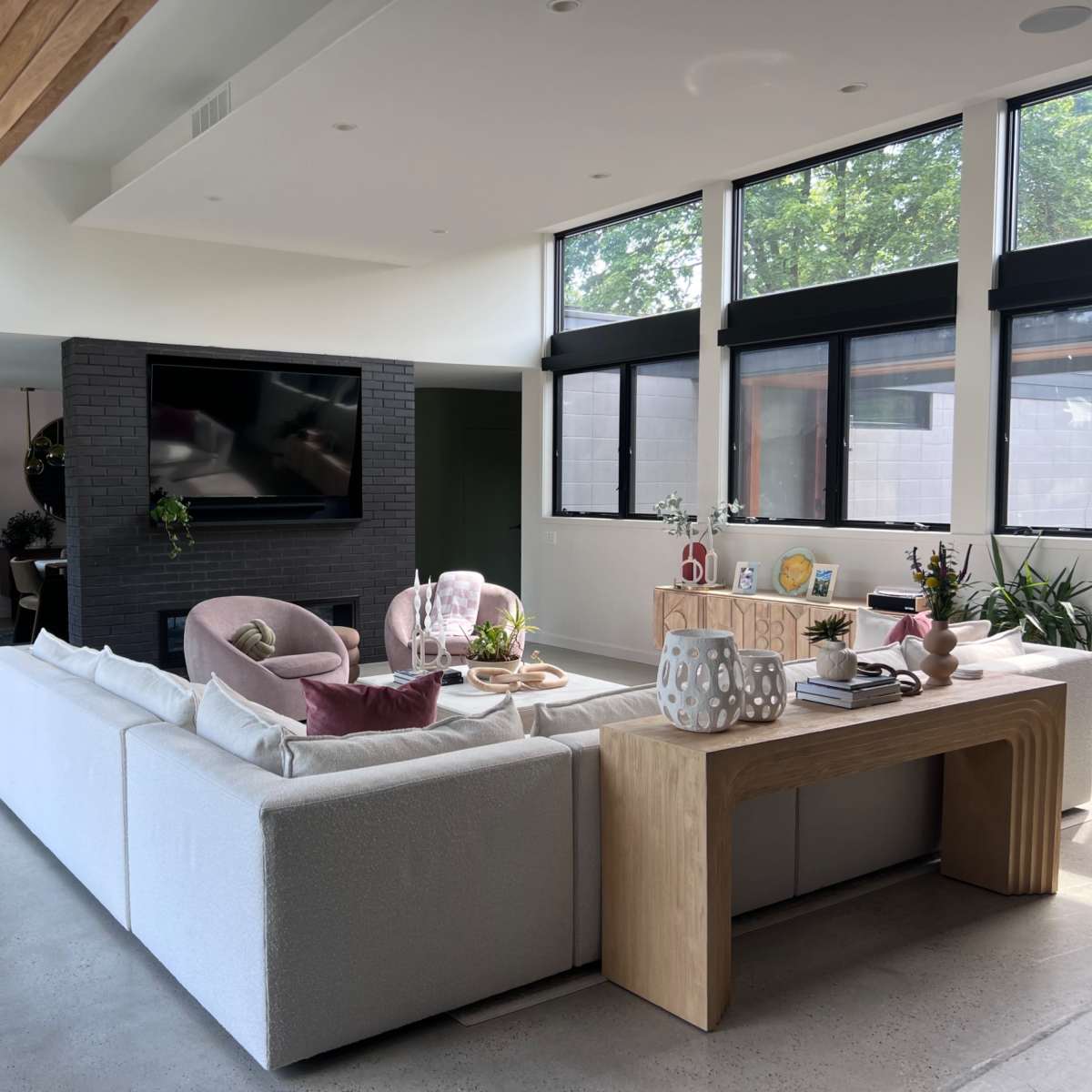 A modern living room with a sectional sofa, a wooden coffee table, large windows letting in natural light, and various plants throughout the space.