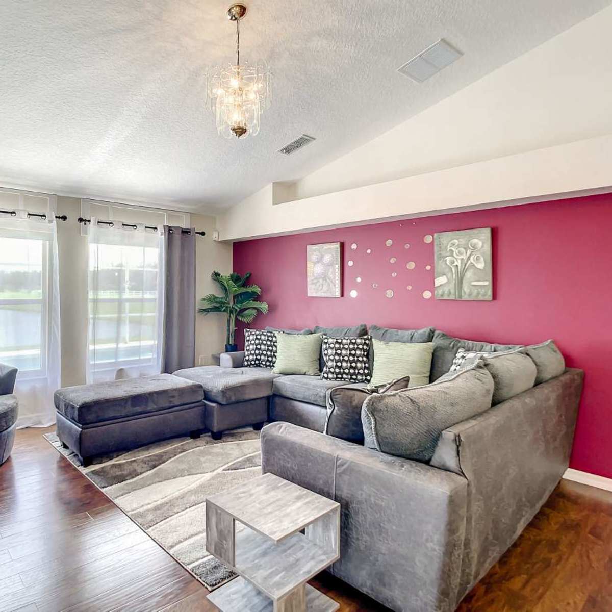 The room features a sectional sofa, an armchair, and large windows with sheer curtains, all arranged around a decorative wall in a blend of colors.