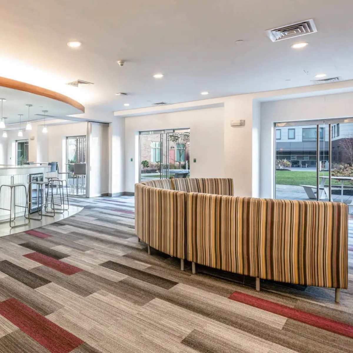 A modern common area with a striped sofa, a bar-style counter, and a dining area with high stools, all situated on patterned carpet.