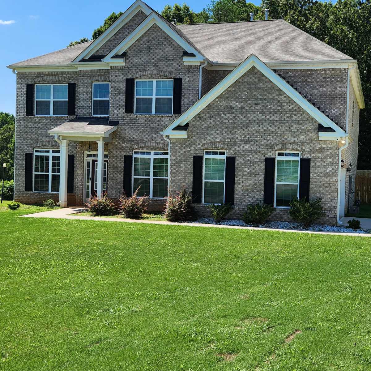 A two-story brick house with a neatly manicured lawn and a paved driveway is situated in a residential area.