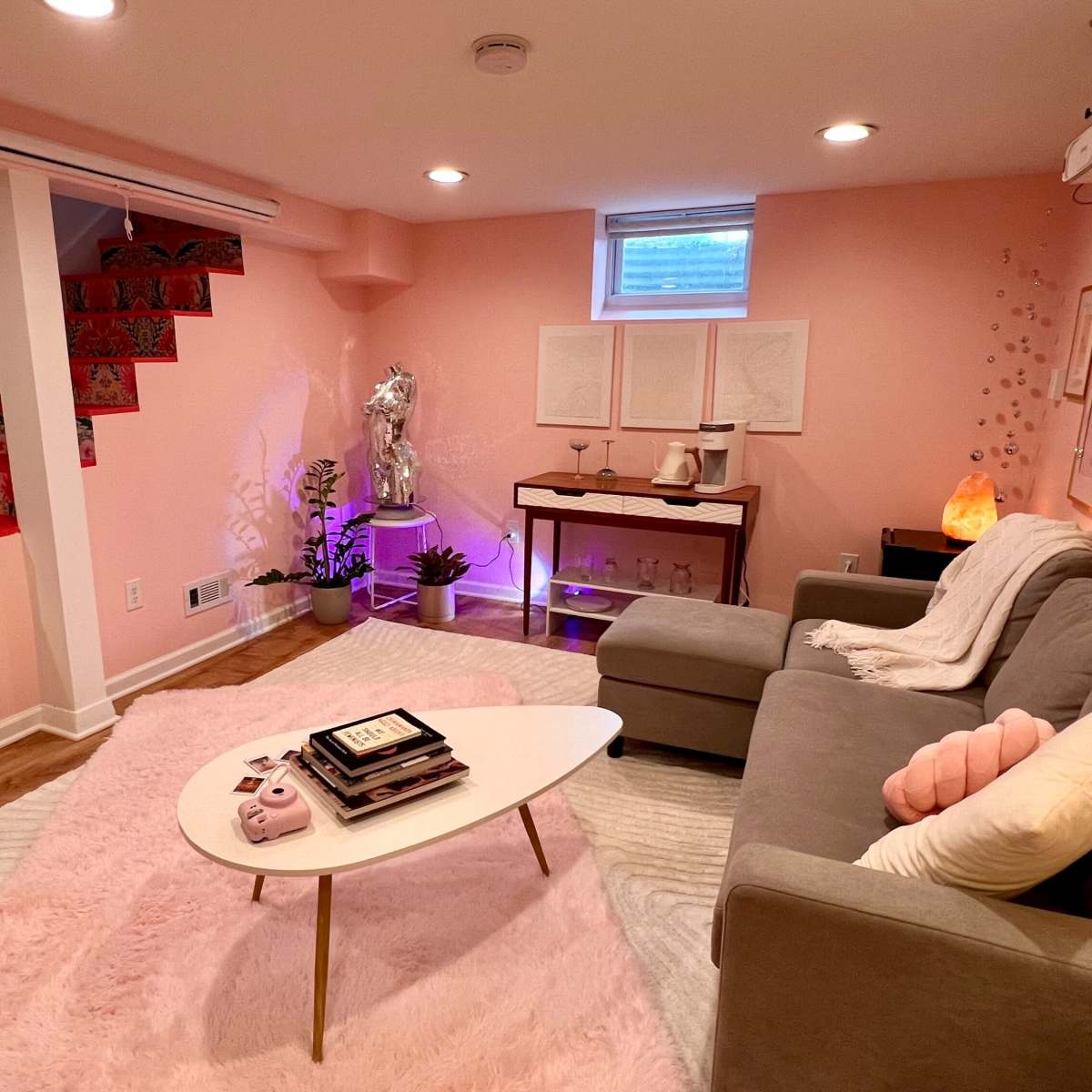 A cozy basement living area with a pink color scheme, featuring a gray sofa, a coffee table, decorative art on the walls, and a staircase with patterned steps.