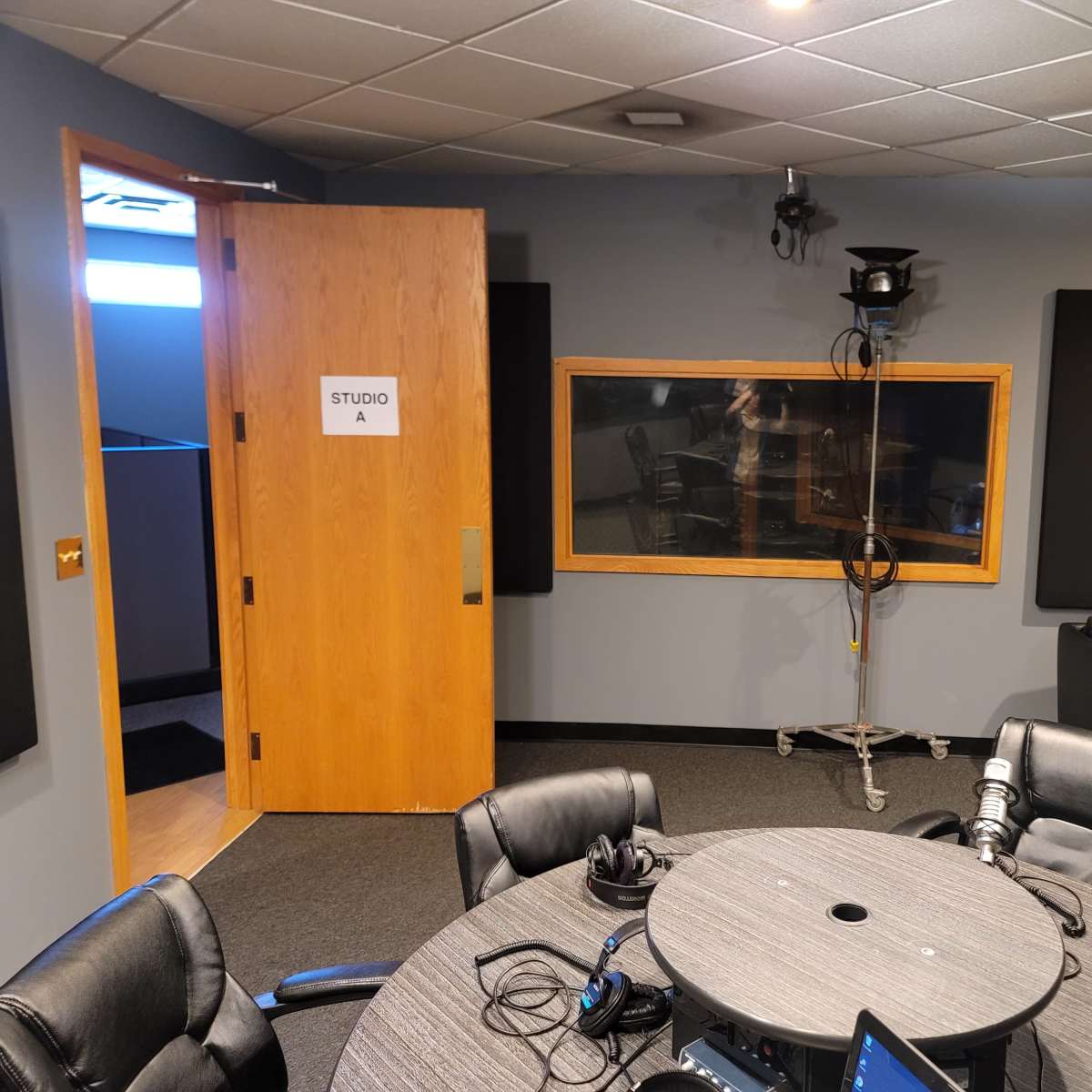 A recording studio with a wooden door labeled "STUDIO A," a circular table surrounded by black chairs, and soundproofing panels on the walls.