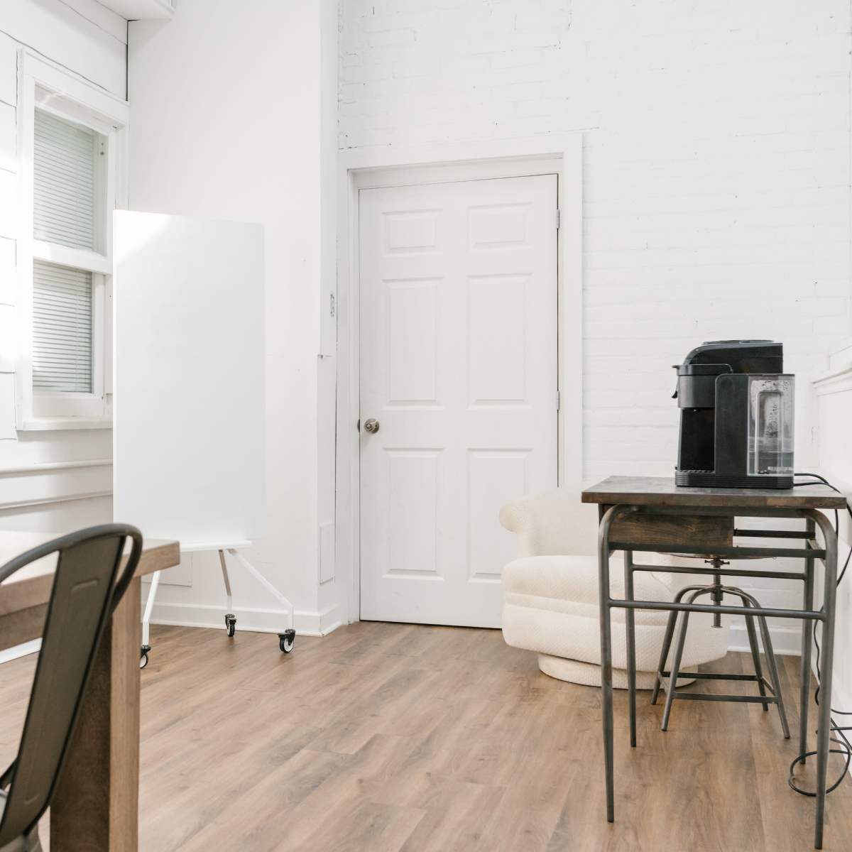 A bright, minimalist office space featuring a wooden table, metal chairs, a whiteboard on wheels, a coffee maker, and a white door against a light-colored wall.