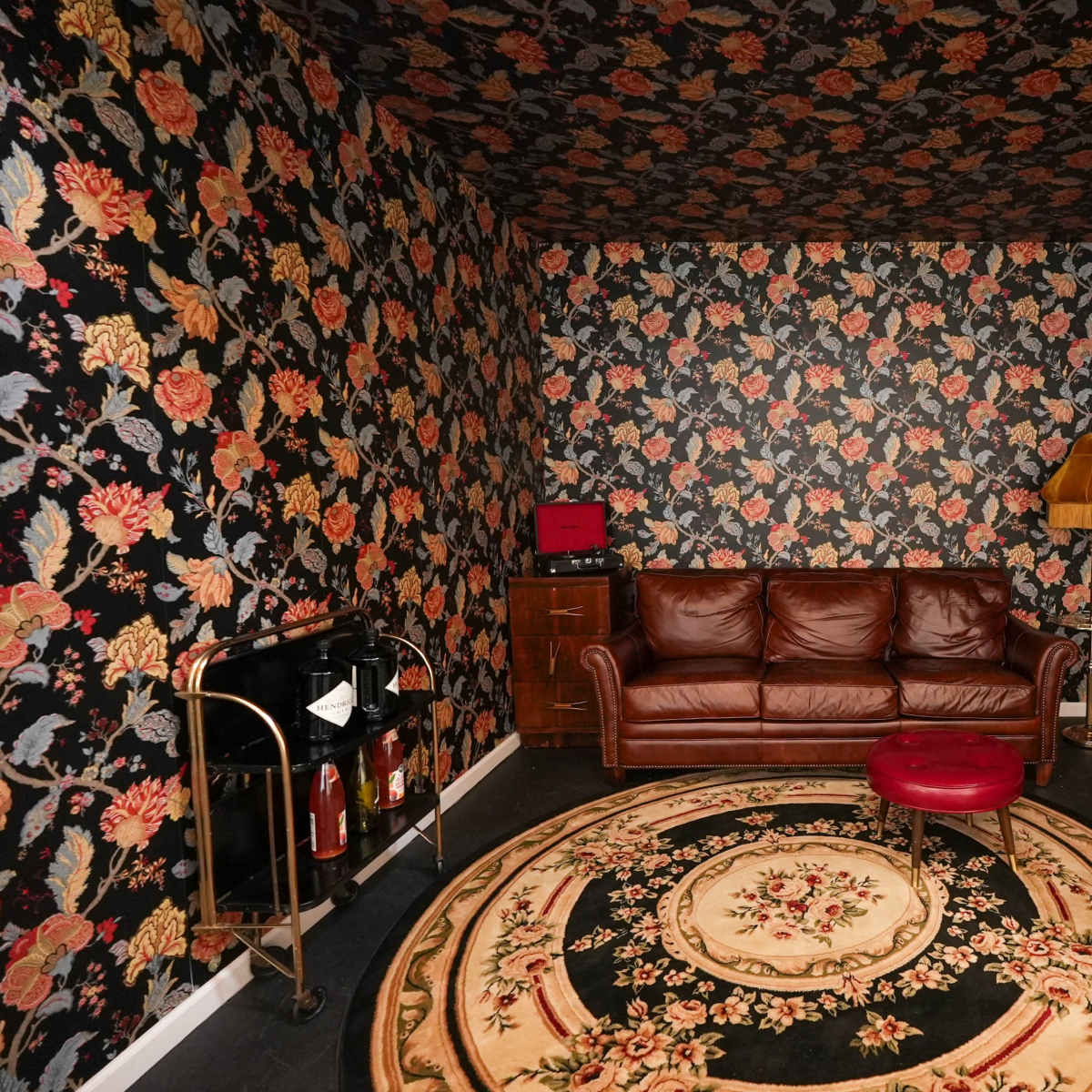 A room with floral wallpaper, a brown leather couch, a circular rug, a wooden dresser, and a bar cart.